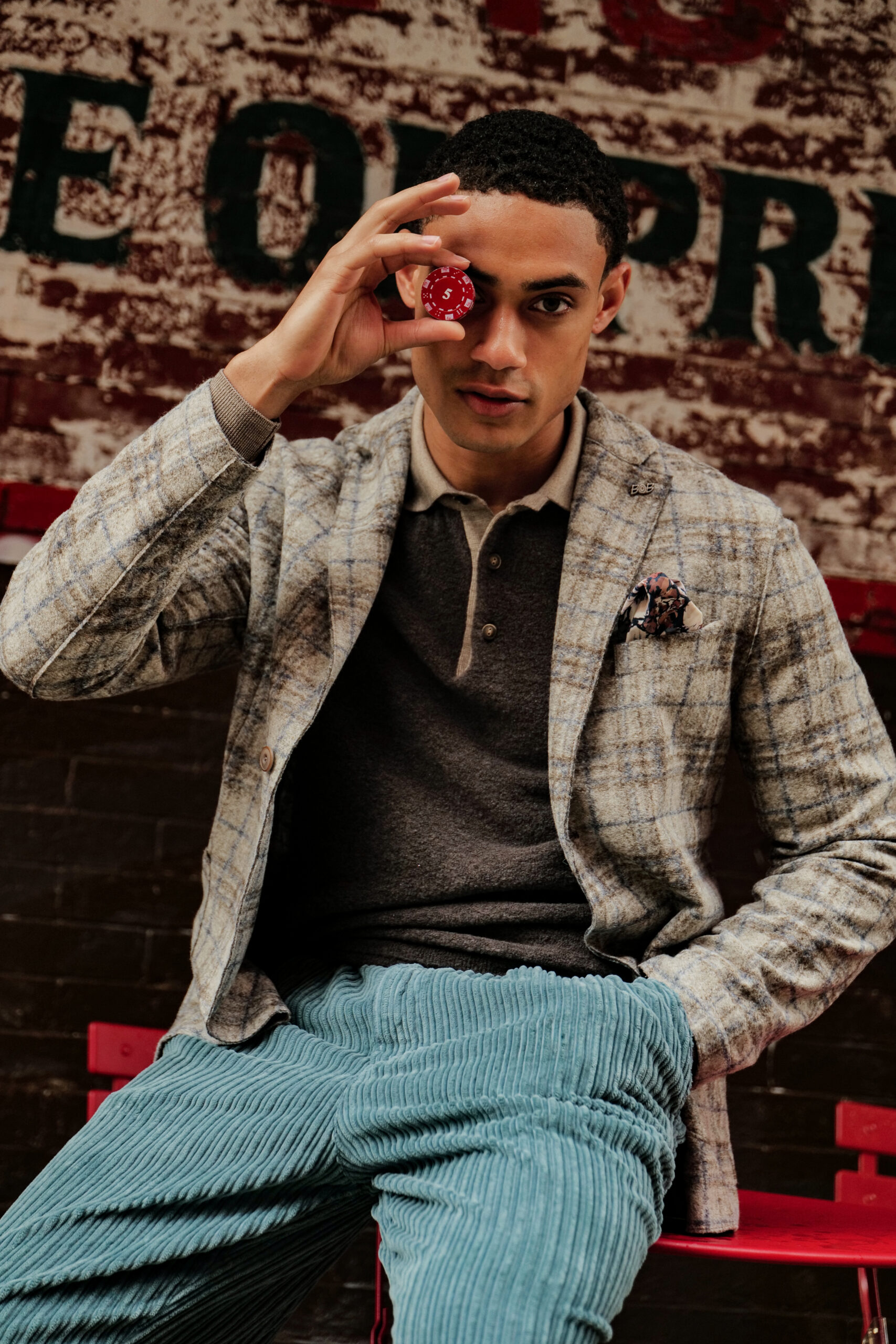 bob-herenmode-blazer-poker Man in een beige geruite colbert van het merk BOB die een rode pokerfiche voor zijn oog houdt